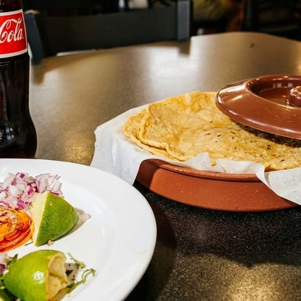A Coca-Cola bottle, a plate of chopped red onions and limes, and a basket of tortillas.
