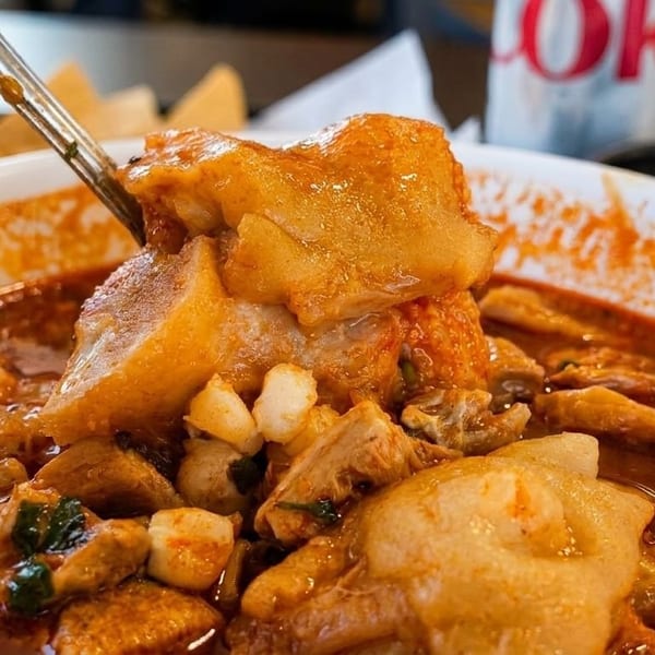 Close-up of a spoon lifting a large piece of meat from a bowl of red menudo soup.
