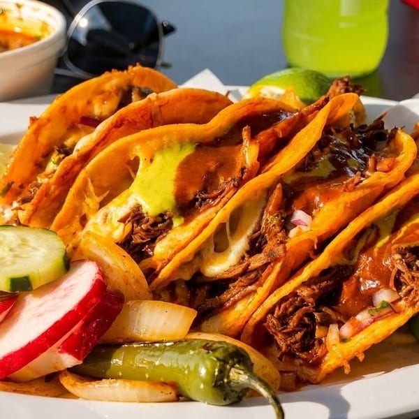 A plate of birria tacos with melted cheese, lime, cucumbers, radishes, and a side of consommé.