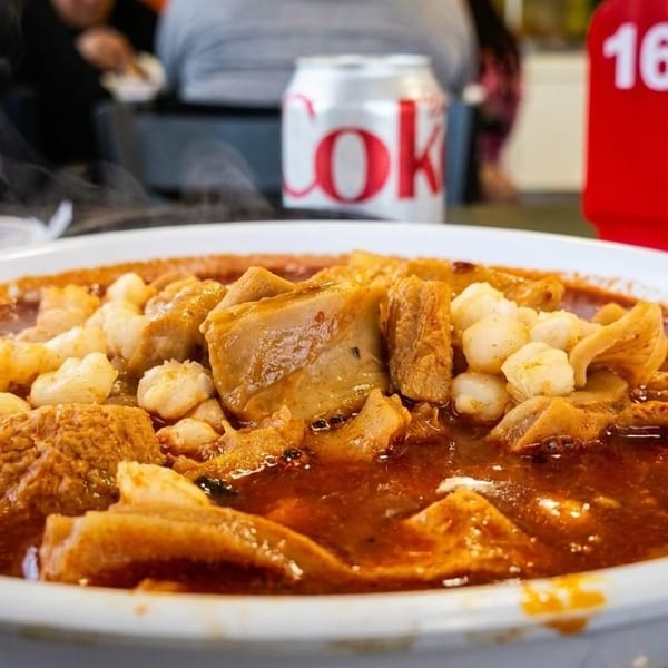 Steaming bowl of menudo with hominy and tripe, a Coke can and table number 16 in the background.