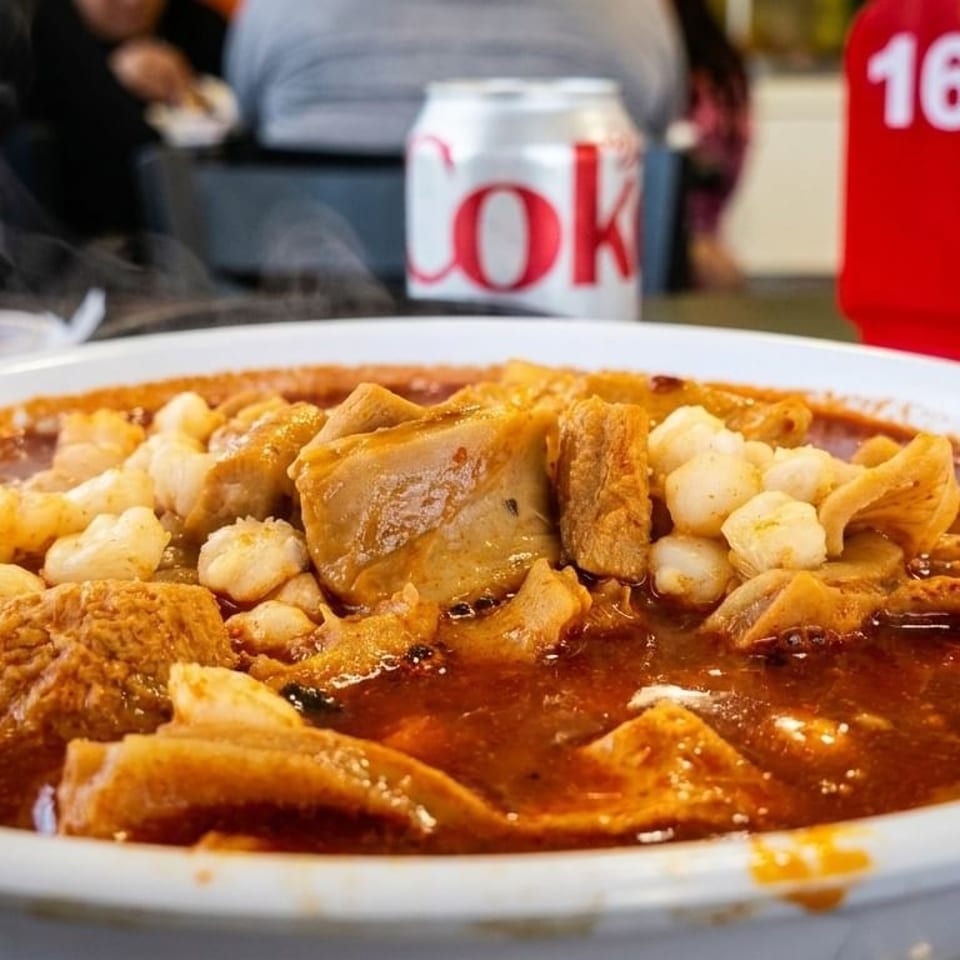 Steaming bowl of menudo with hominy and tripe, a Coke can and table number 16 in the background.