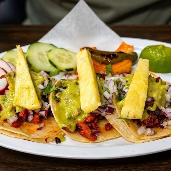 Three al pastor tacos with pineapple, guacamole, and red onion, served with radishes, cucumbers, and limes.