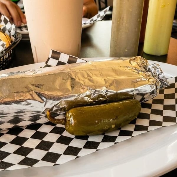 Foil-wrapped burrito with a whole pickled jalapeño on a checkered paper-lined plate.