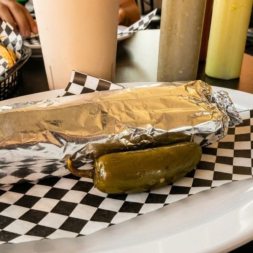 Foil-wrapped burrito with a whole pickled jalapeño on a checkered paper-lined plate.