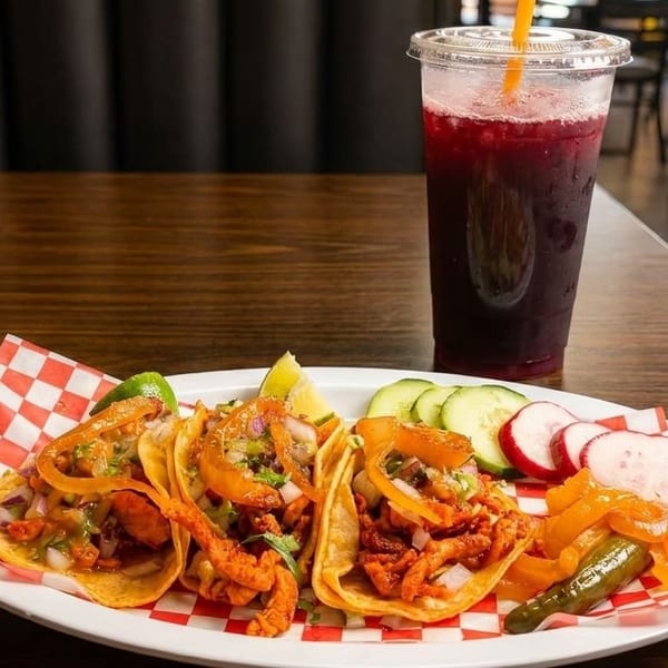 Three tacos with meat, onions, and lime, served with a drink, cucumbers, and radishes.
